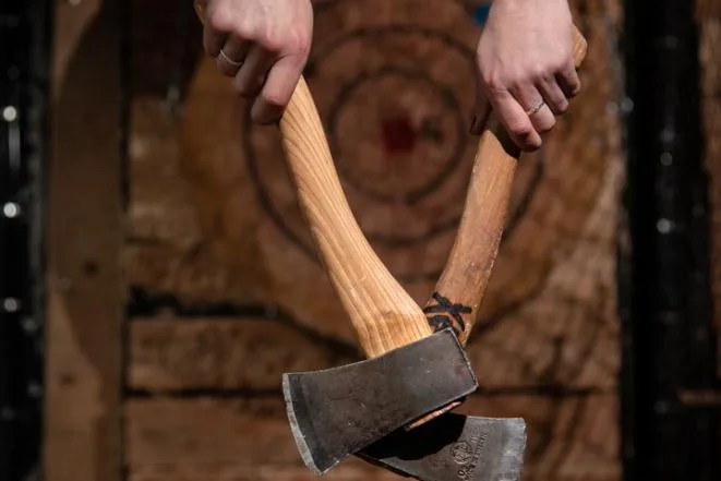 Two hands holding throwing axes in front of a wooden target